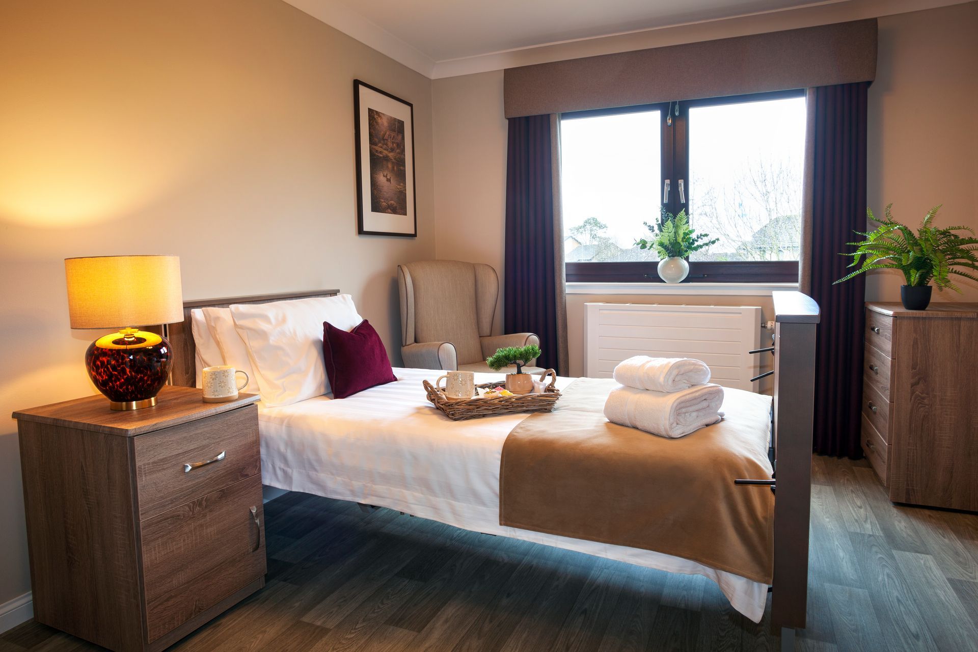 A patient room with a single bed, dresser, window, and armchair. Light brown walls and curtains.
