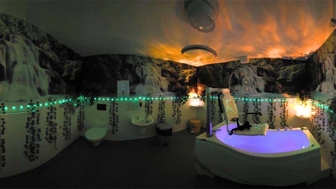 Fantasy-themed bathroom with waterfall wallpaper, a tub, and hanging green lights; lit with orange and purple hues.