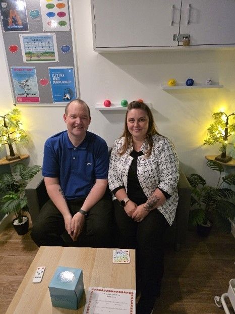 Two people seated on a sofa. One wears a blue shirt, the other a black top and white patterned jacket. They are in a room with plants.