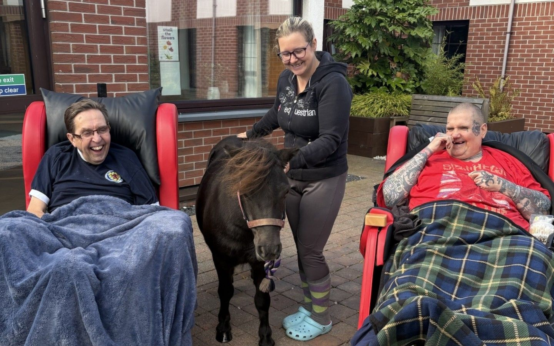 Two men in wheelchairs smile as a woman stands with a small pony outside a building.