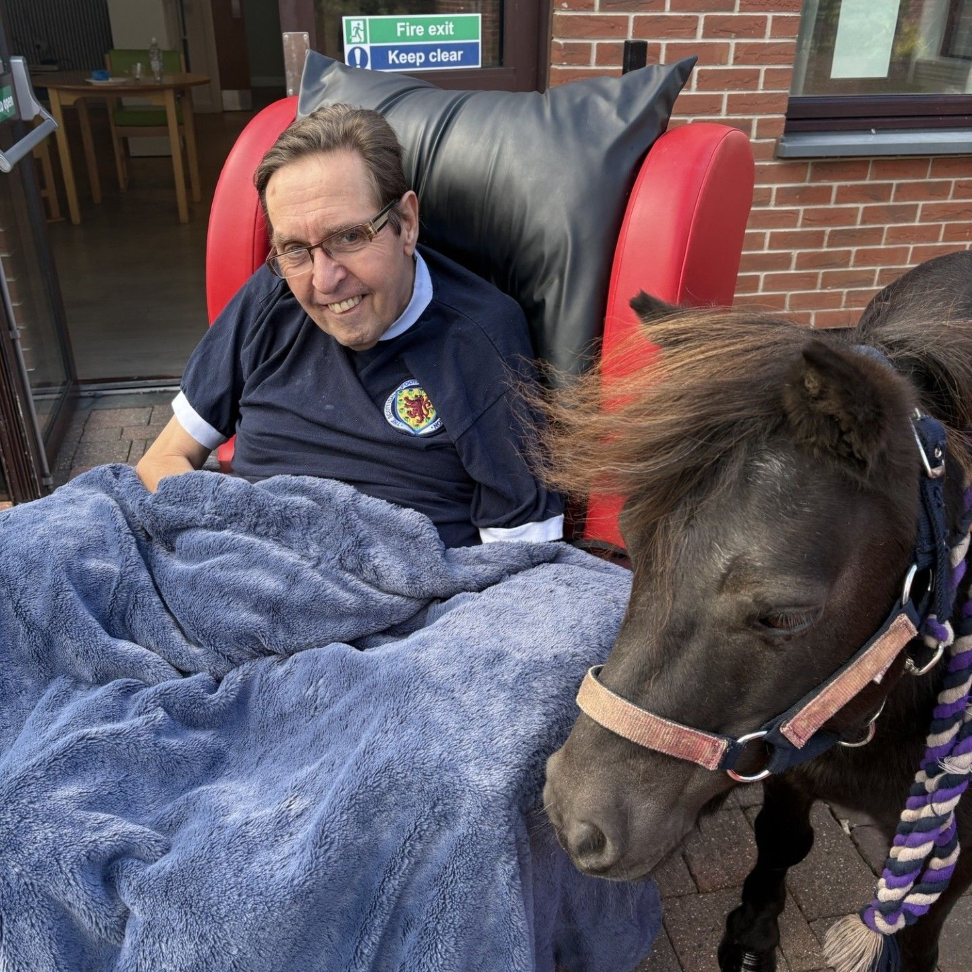 Man in red chair with pony; man smiles, wrapped in blue blanket. Pony wears halter, next to man outside.