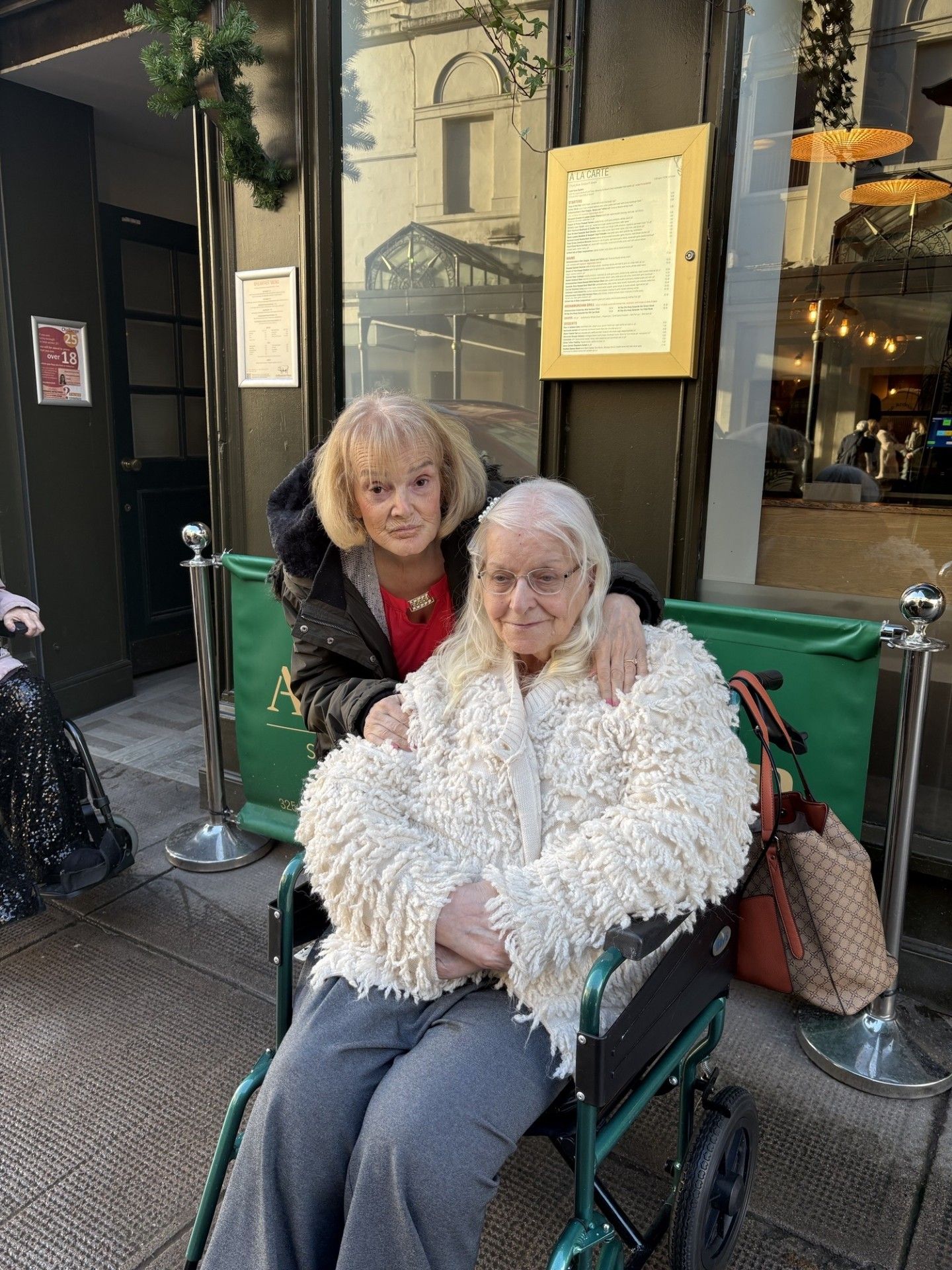 Woman in a wheelchair with another woman's arm around her, smiling outside a restaurant.