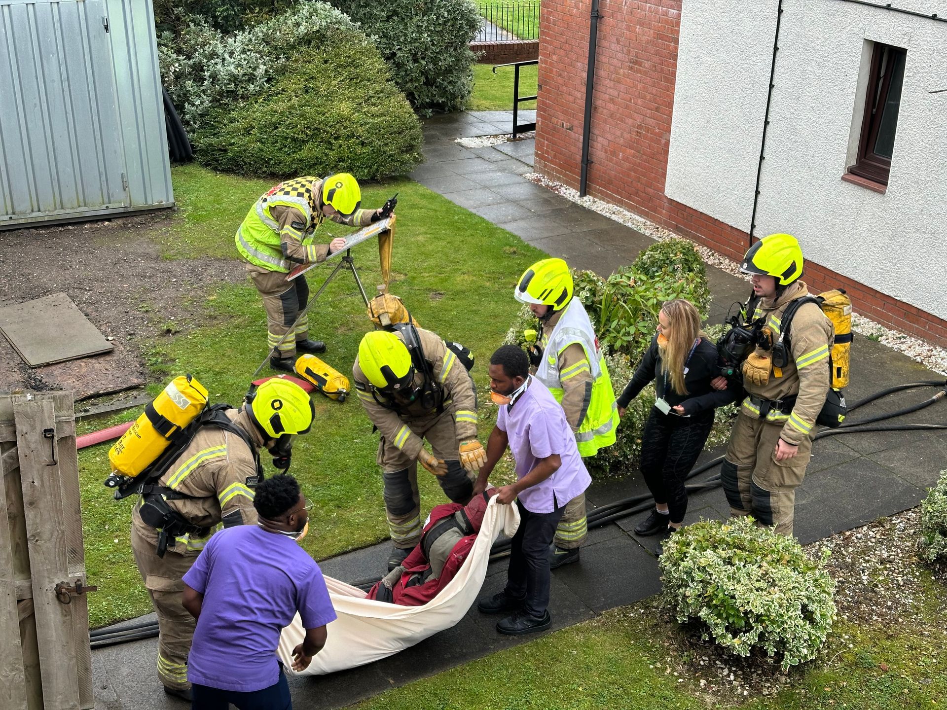 Firefighters carry a dummy on a stretcher. Rescuers, wearing helmets and gear, are in a grassy area beside a building.