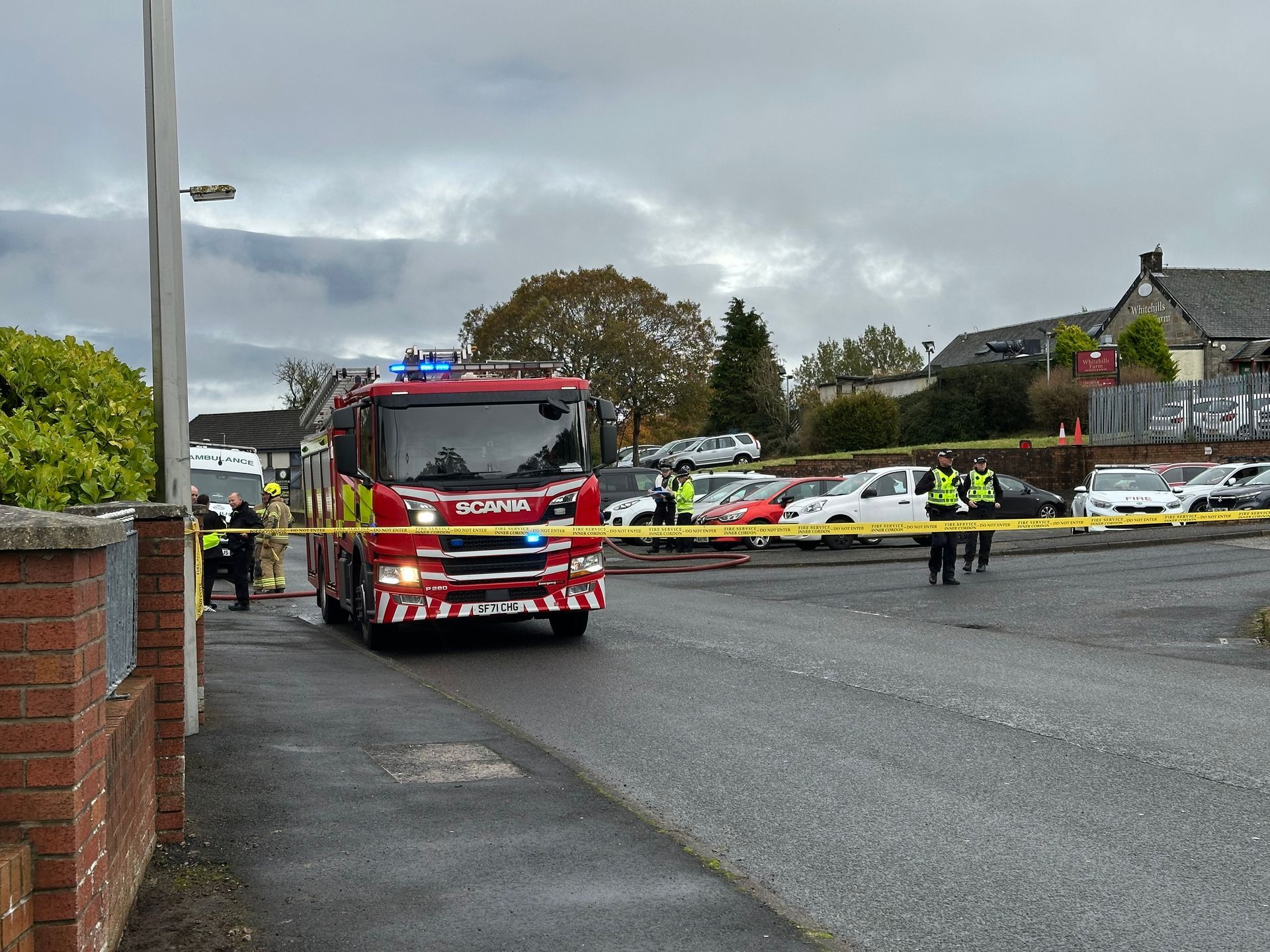 Fire truck at a scene, cordoned off. Emergency personnel and cars are visible in the background on an overcast day.