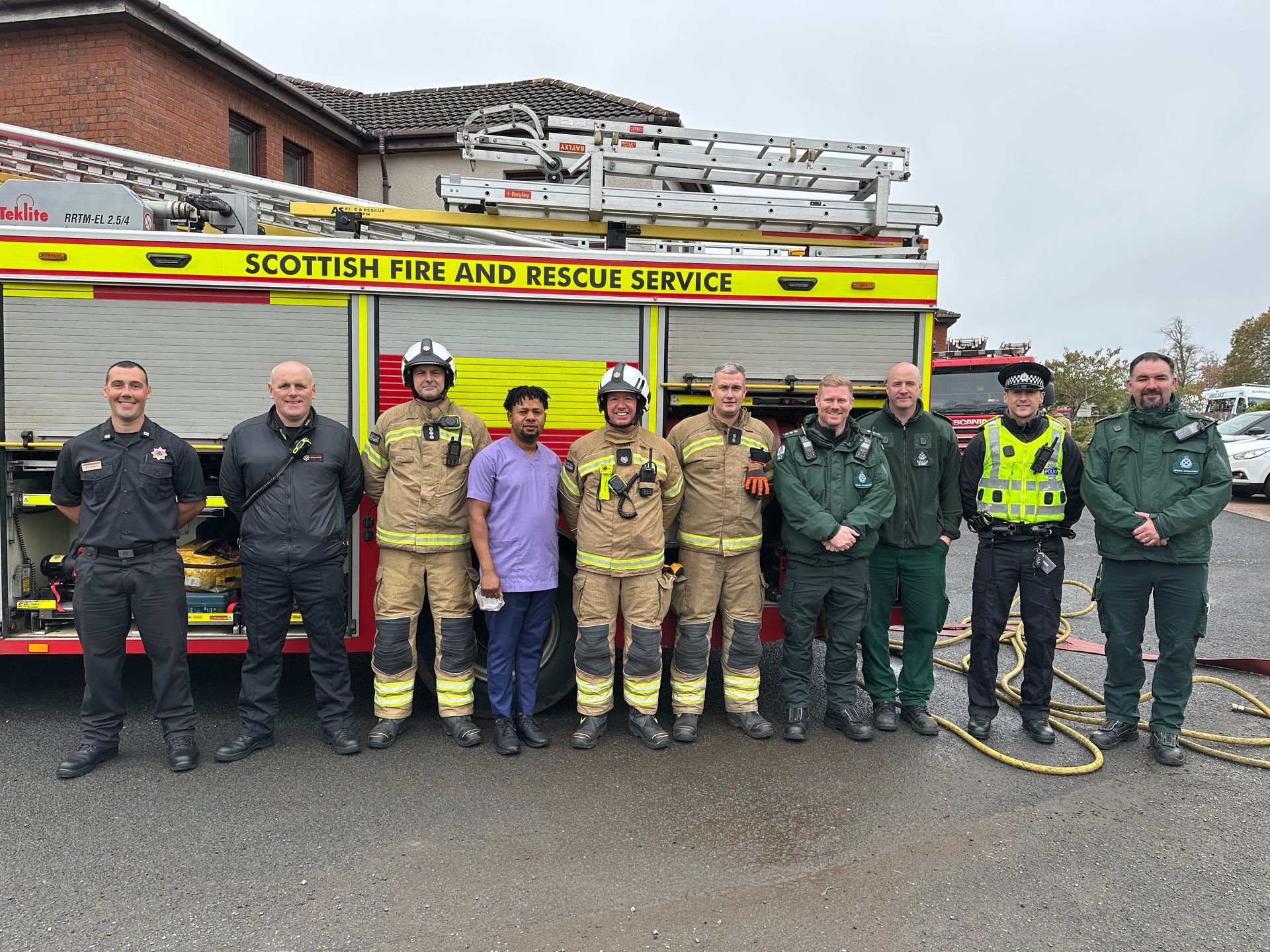 Firefighters, medics, and police stand in front of a fire truck. They are smiling and posing outdoors.