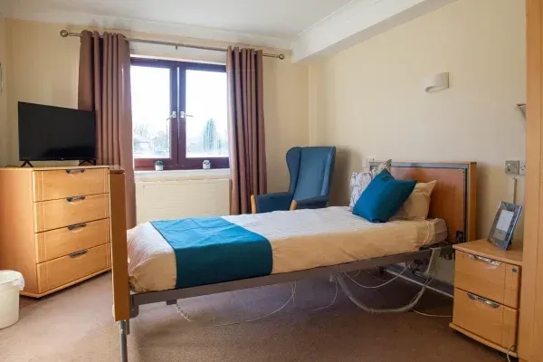 A patient room with a single bed, dresser, window, and armchair. Light brown walls and curtains.