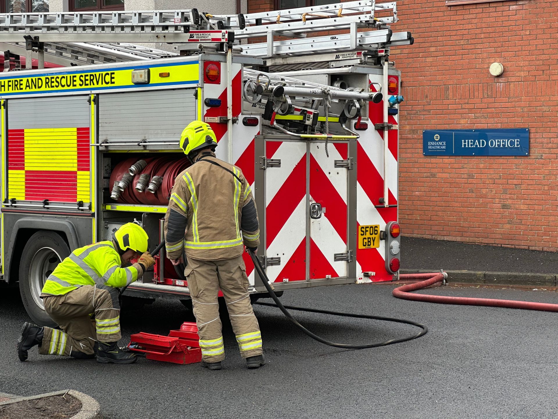Firefighters near a fire truck, preparing equipment. Exterior setting with brick building.