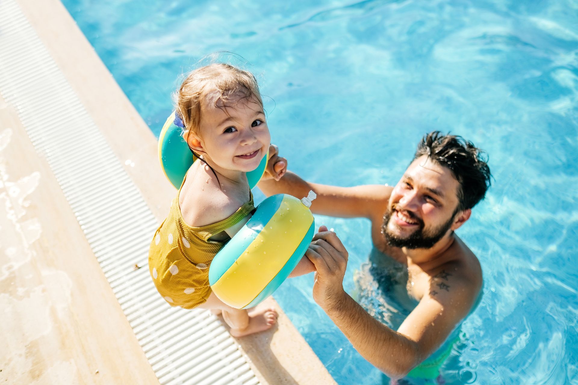 Child with floaties smiling in a swimming pool with an adult nearby