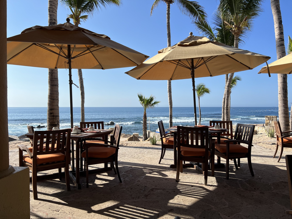 Outdoor beachside restaurant with tables, chairs, and umbrellas; ocean and palm trees visible.