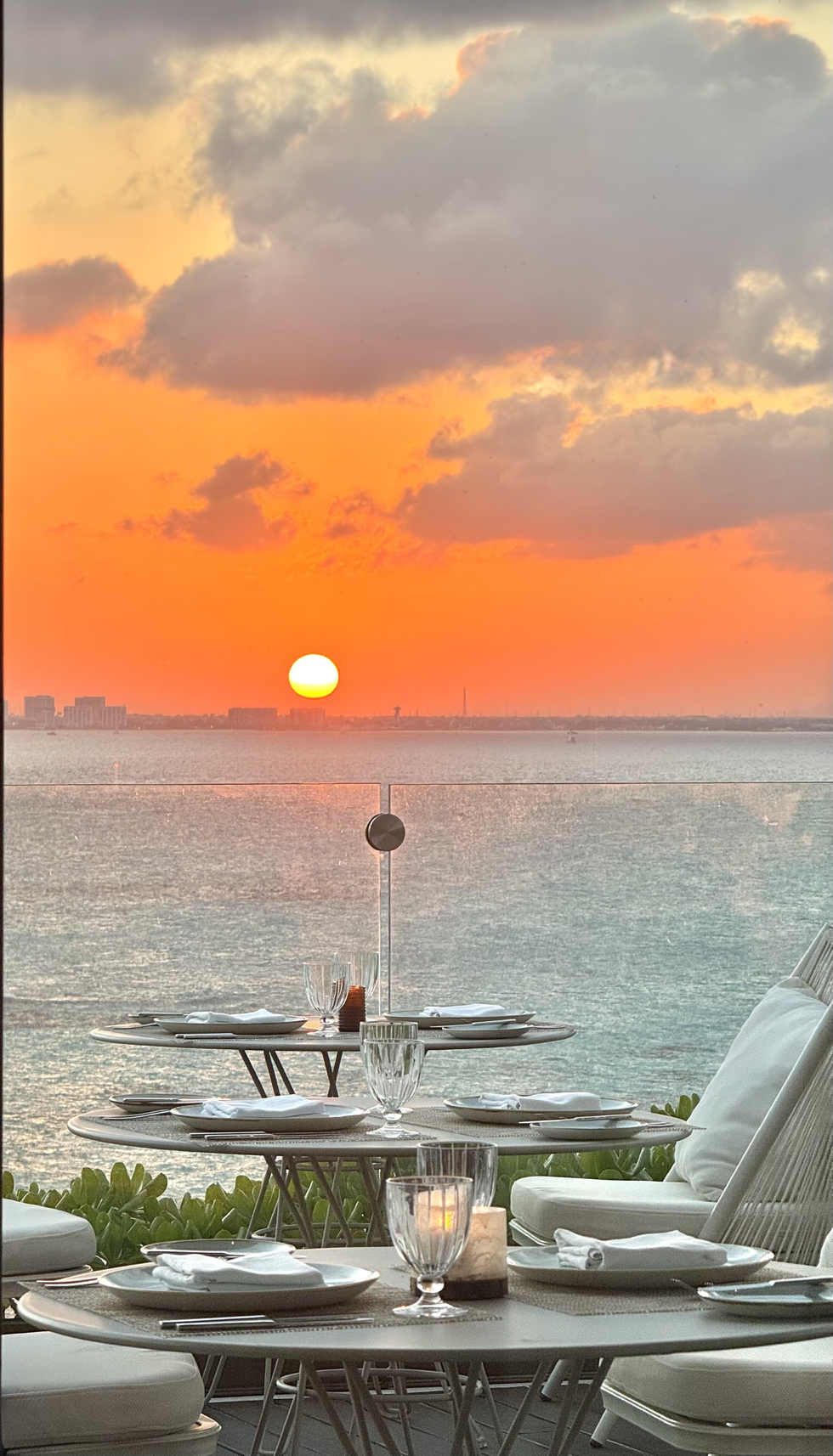Oceanfront restaurant tables set for dinner with a sunset backdrop: orange sky, sea, and city silhouette.