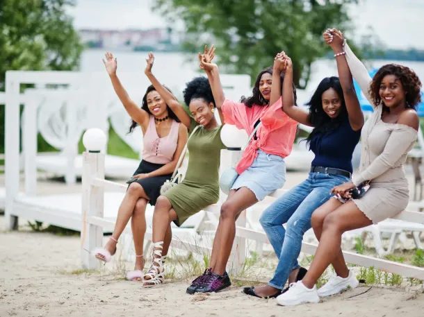 Five women sitting on a white fence, raising their arms in celebration. Beach setting.