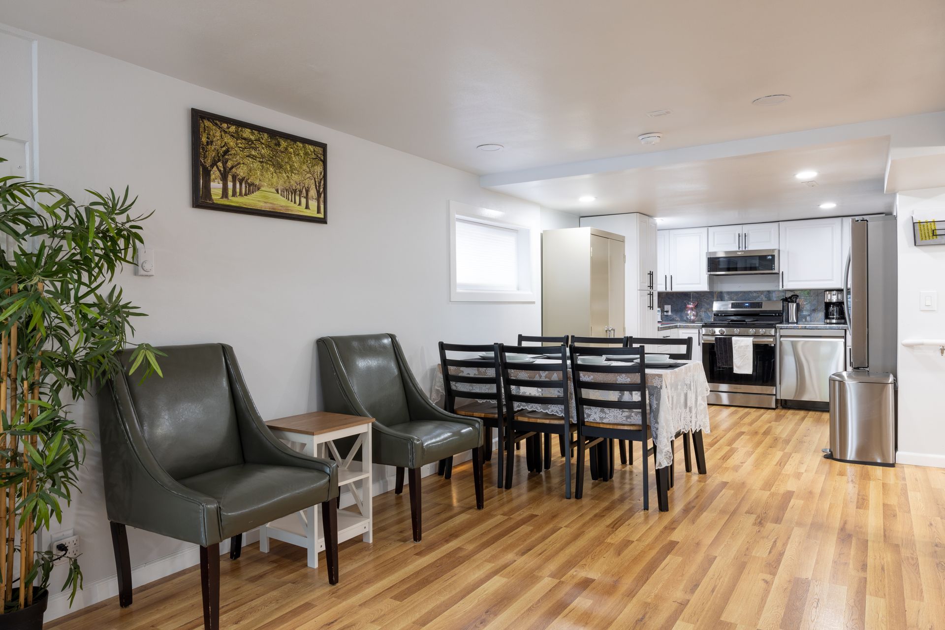 Dining area with chairs, table, kitchen in the background, wood floors, and decorative plants.