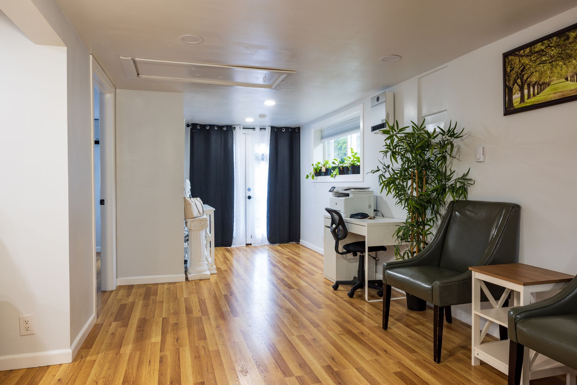 A bright interior room with wood floors, white walls, and a desk. Two chairs and a doorway are visible.