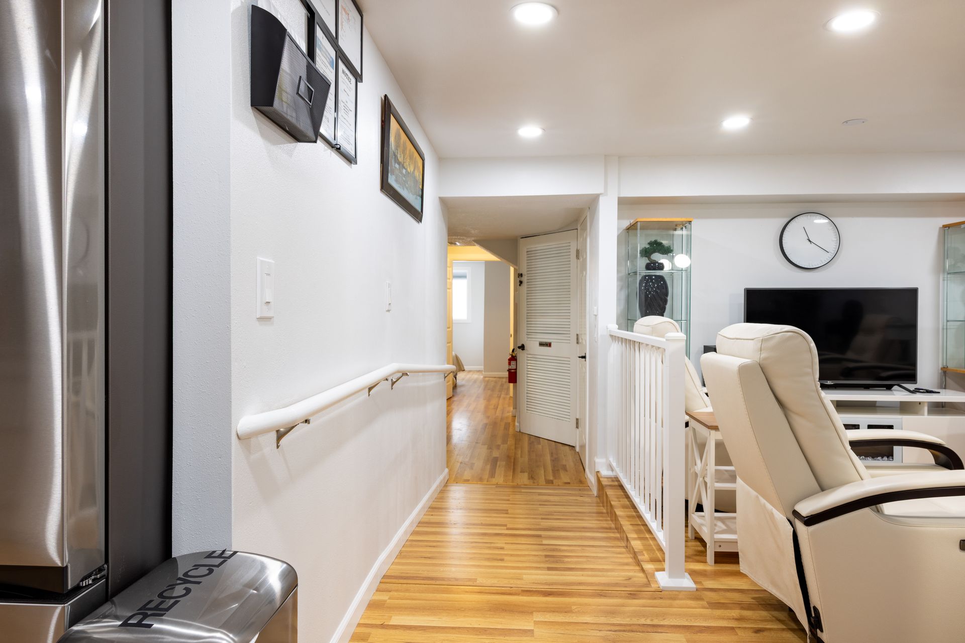 Hallway with hardwood floor, handrail, and white walls leads to an open living area with a TV and recliner.