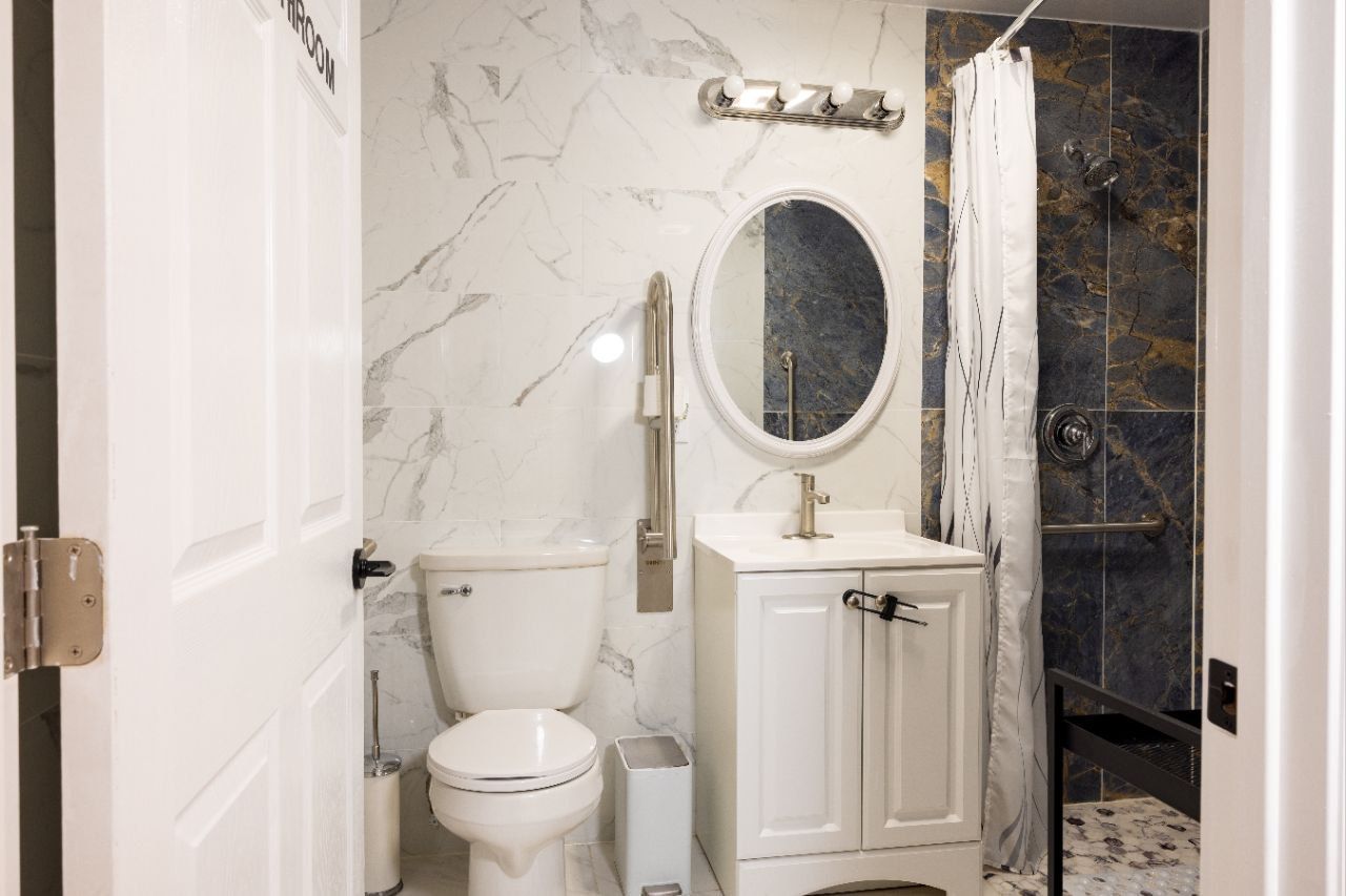 Small, white-tiled bathroom with toilet, sink, and accessible shower. Marble-look accent wall.
