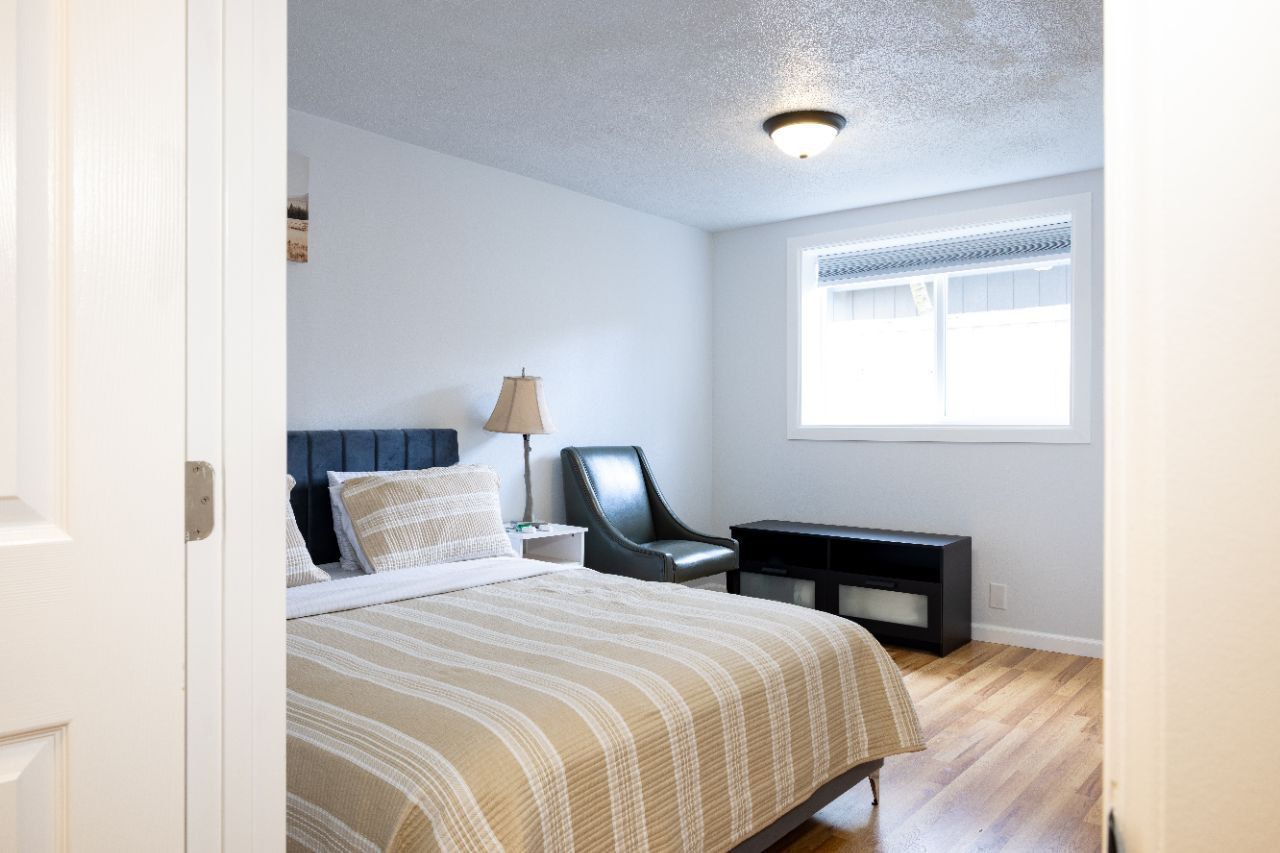 Bedroom interior: bed with blue headboard, armchair, nightstand with lamp, window, and wood floor.