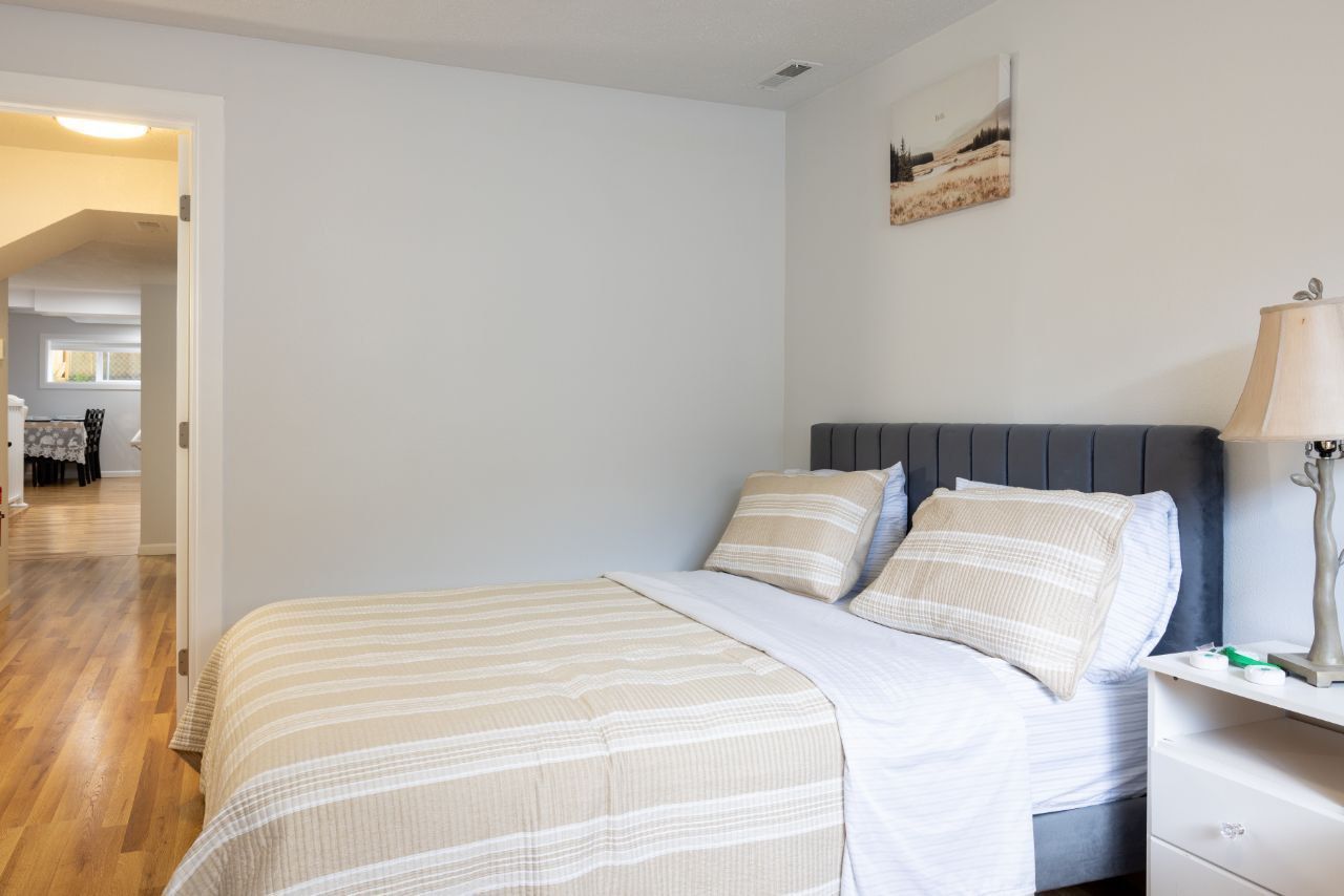 Bedroom with bed, nightstand, lamp, and artwork. A doorway leads to another room with hardwood floors.