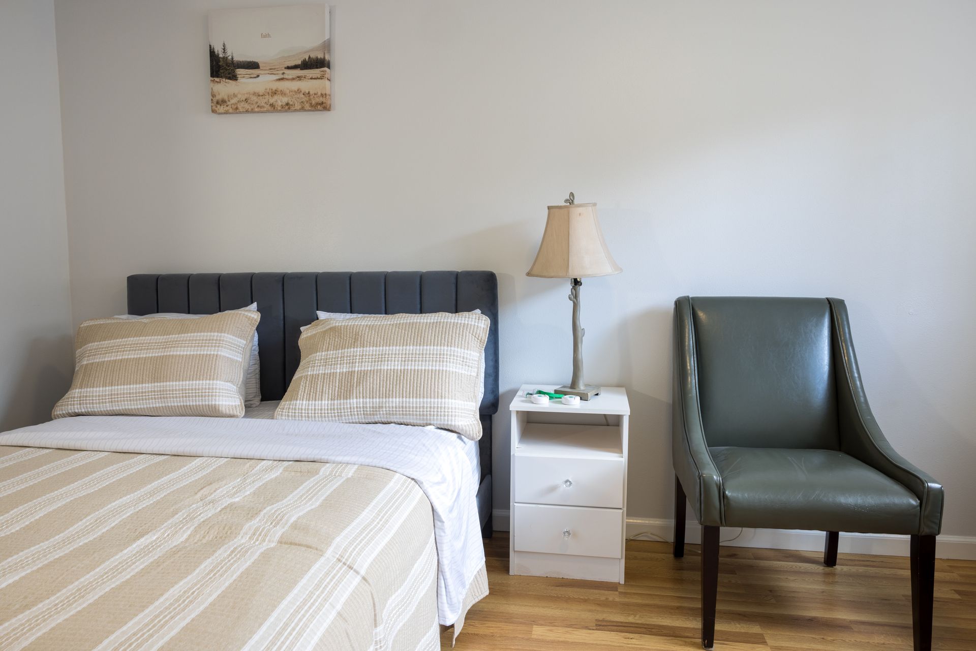 Bedroom with bed, nightstand, lamp, chair, and art on a light gray wall.