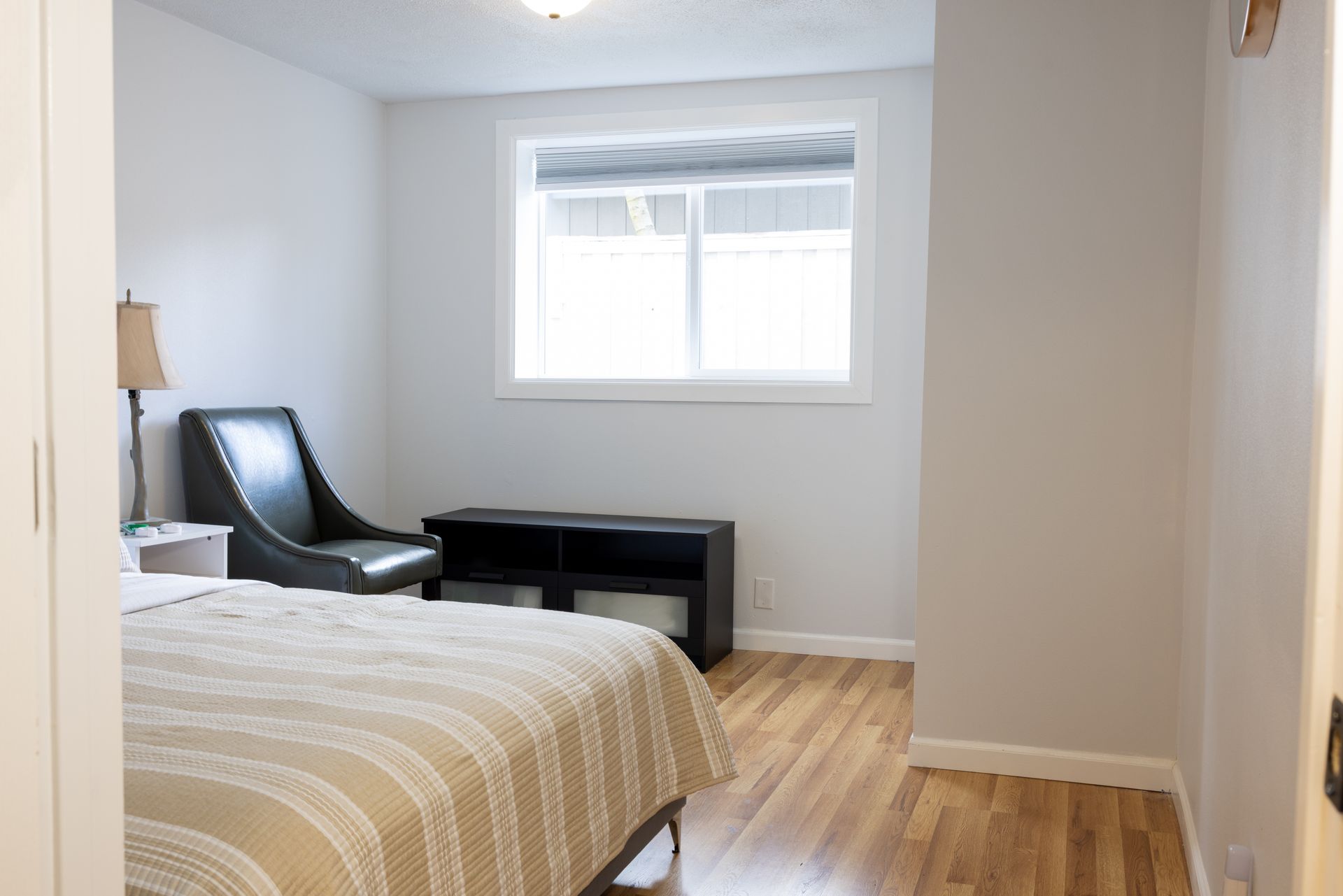 Bedroom with a bed, chair, window, and wooden floor. Neutral colors with natural light.