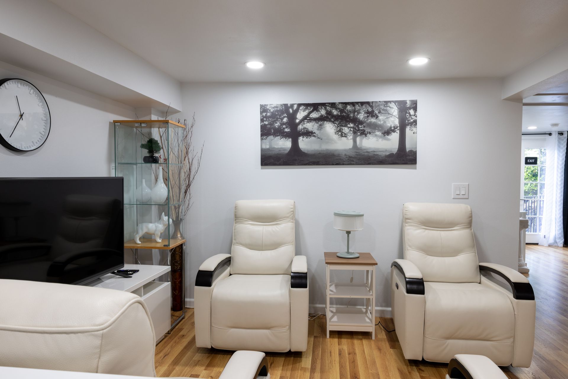 Living room with two cream recliners, a side table, TV, art print, and a glass cabinet. White walls and wood floor.