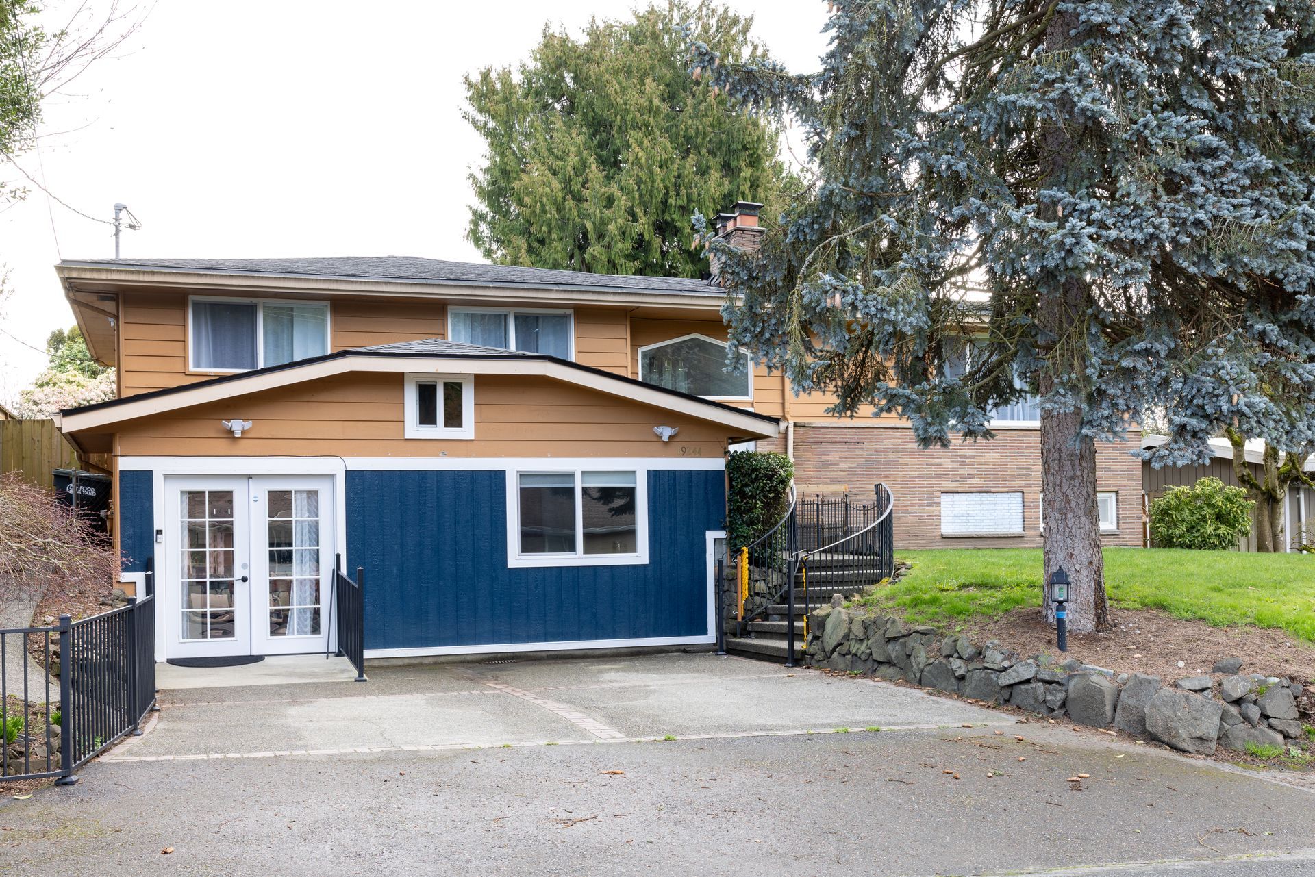 Two-story house with blue and brown siding, a driveway, and a small yard with a large tree.