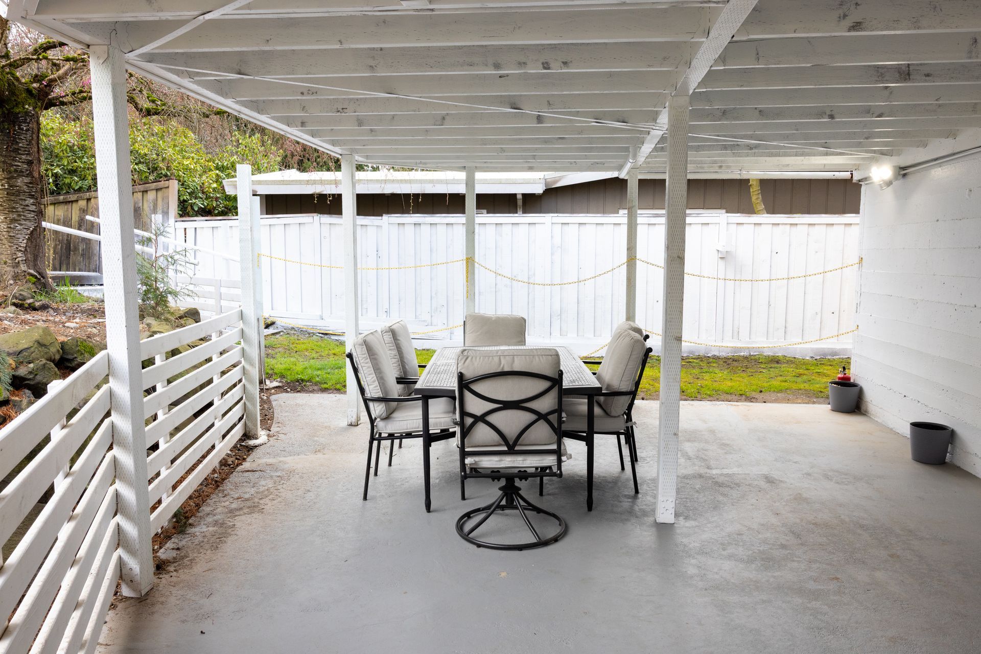Outdoor dining area with a table and chairs under a white-painted pergola, adjacent to a white fence.