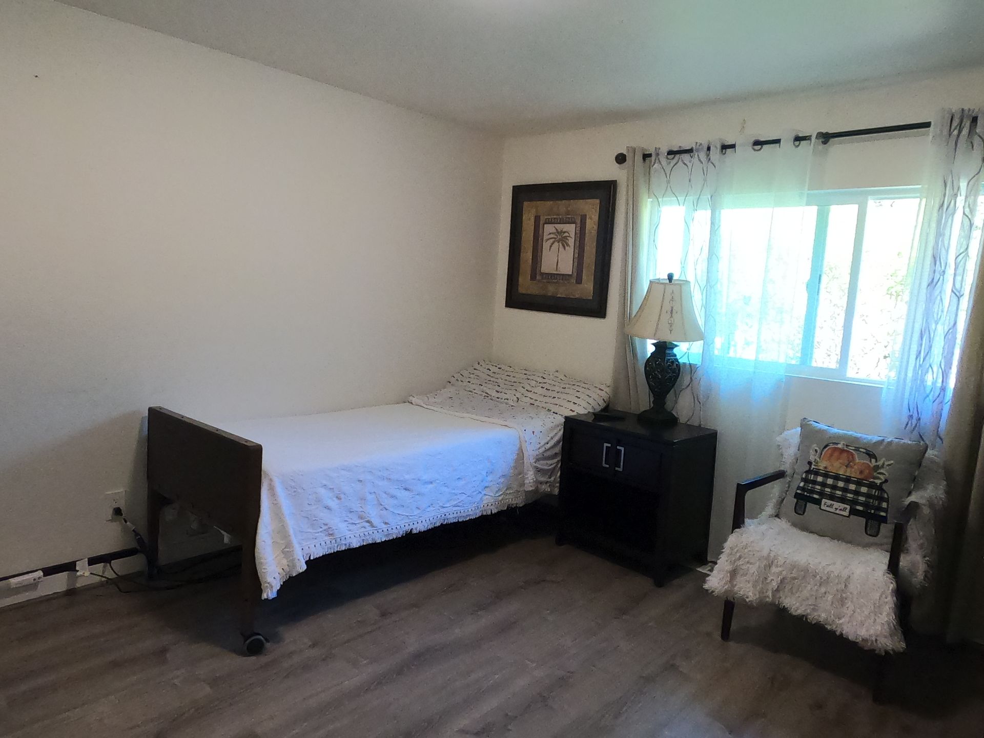 Bedroom with hospital bed, nightstand with lamp, chair by window with fall pillow.