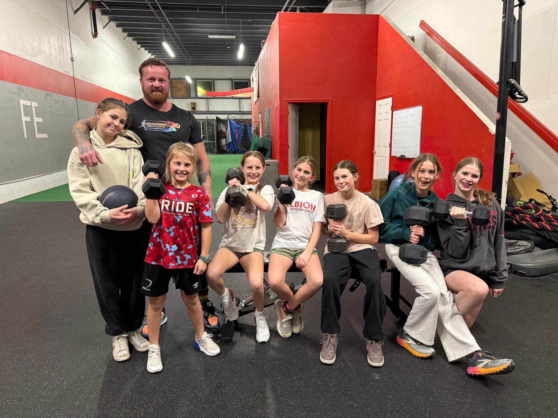 A coach stands with a group of people holding small hand weights in a gym with red walls and a black floor.