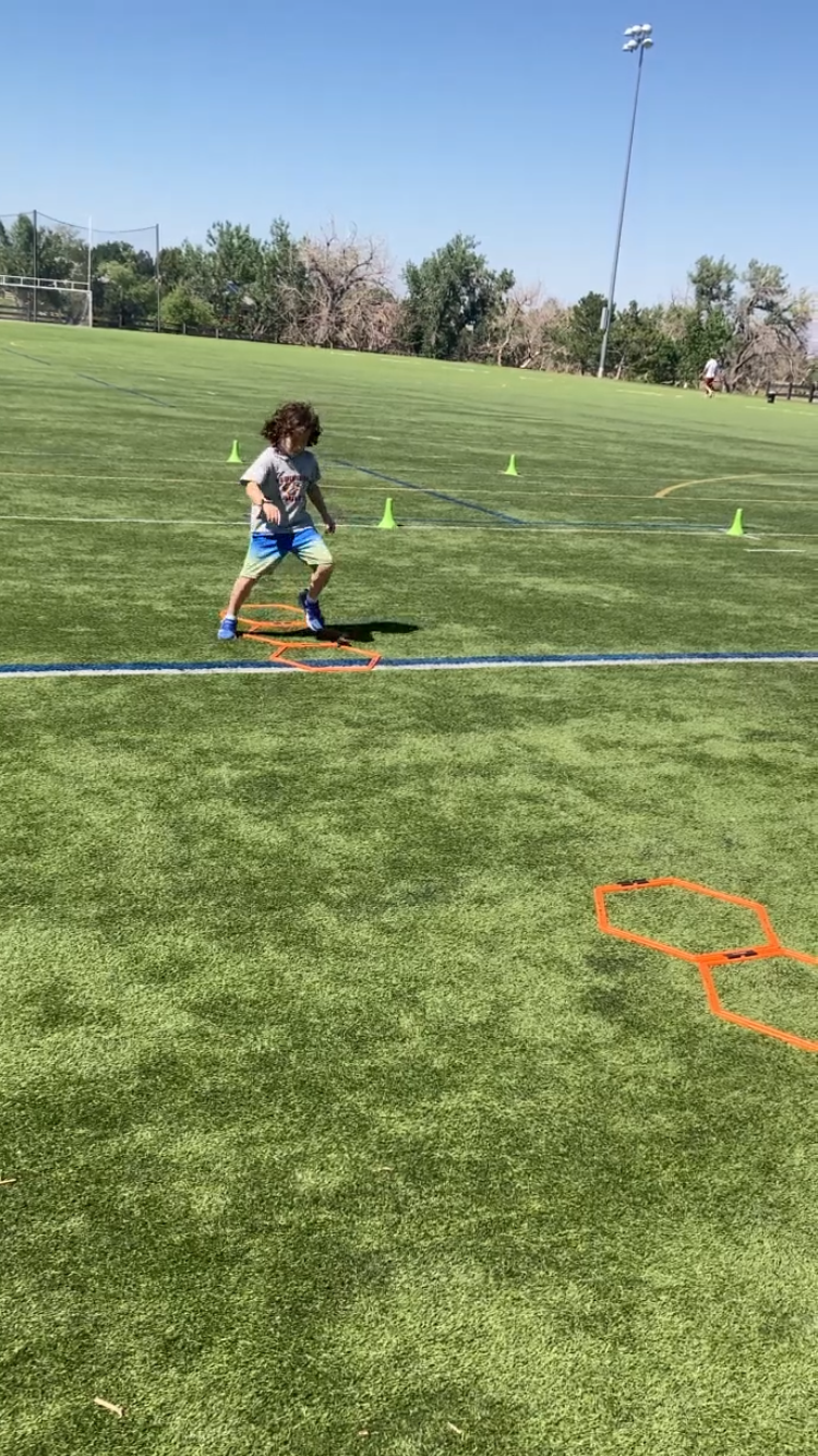 A person in a gray shirt and blue shorts jumping through orange agility hoops on a grass field on a sunny day.