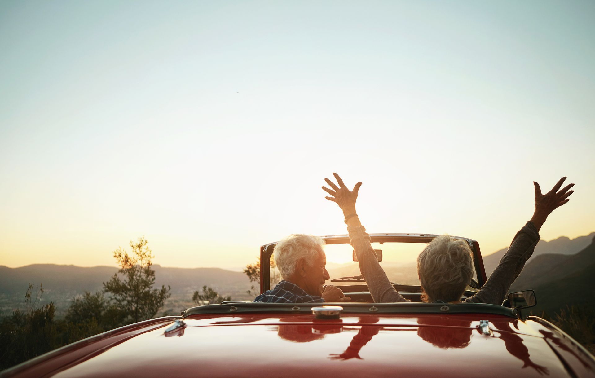 Two people in a red convertible with arms raised, enjoying a sunset view over mountains.