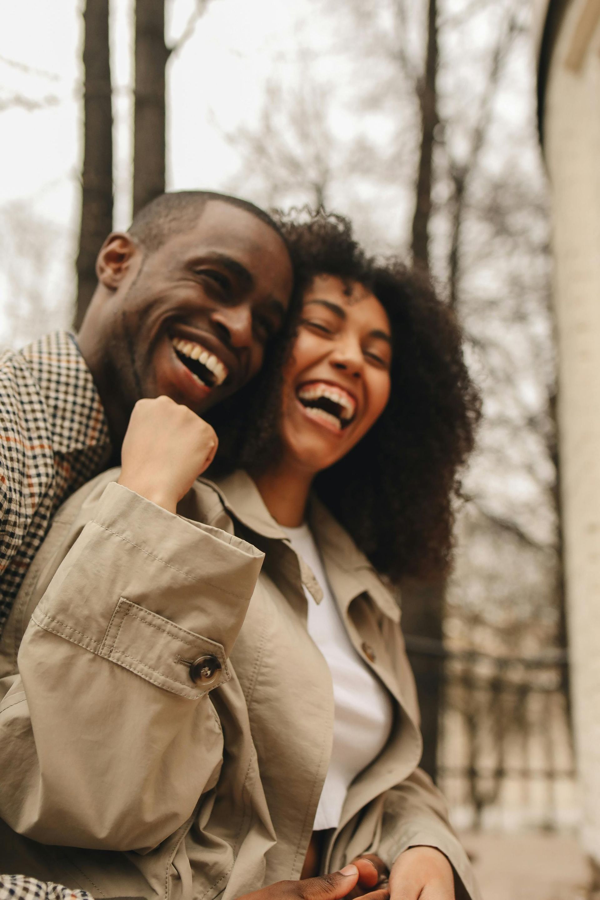Smiling couple embraces outdoors, laughing. Man in patterned coat, woman in tan jacket.
