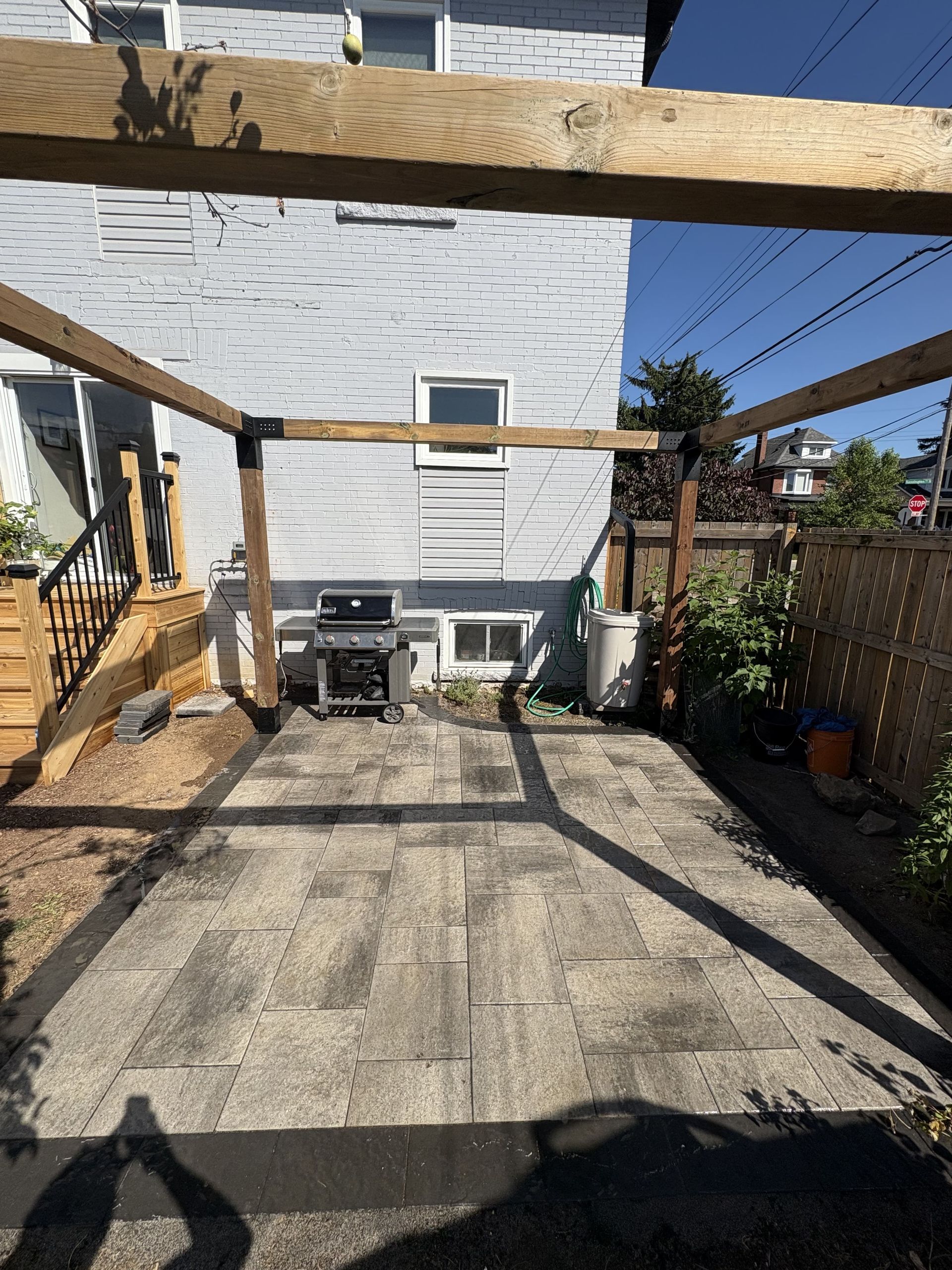 Pergola frames a paved patio with a grill and white building.