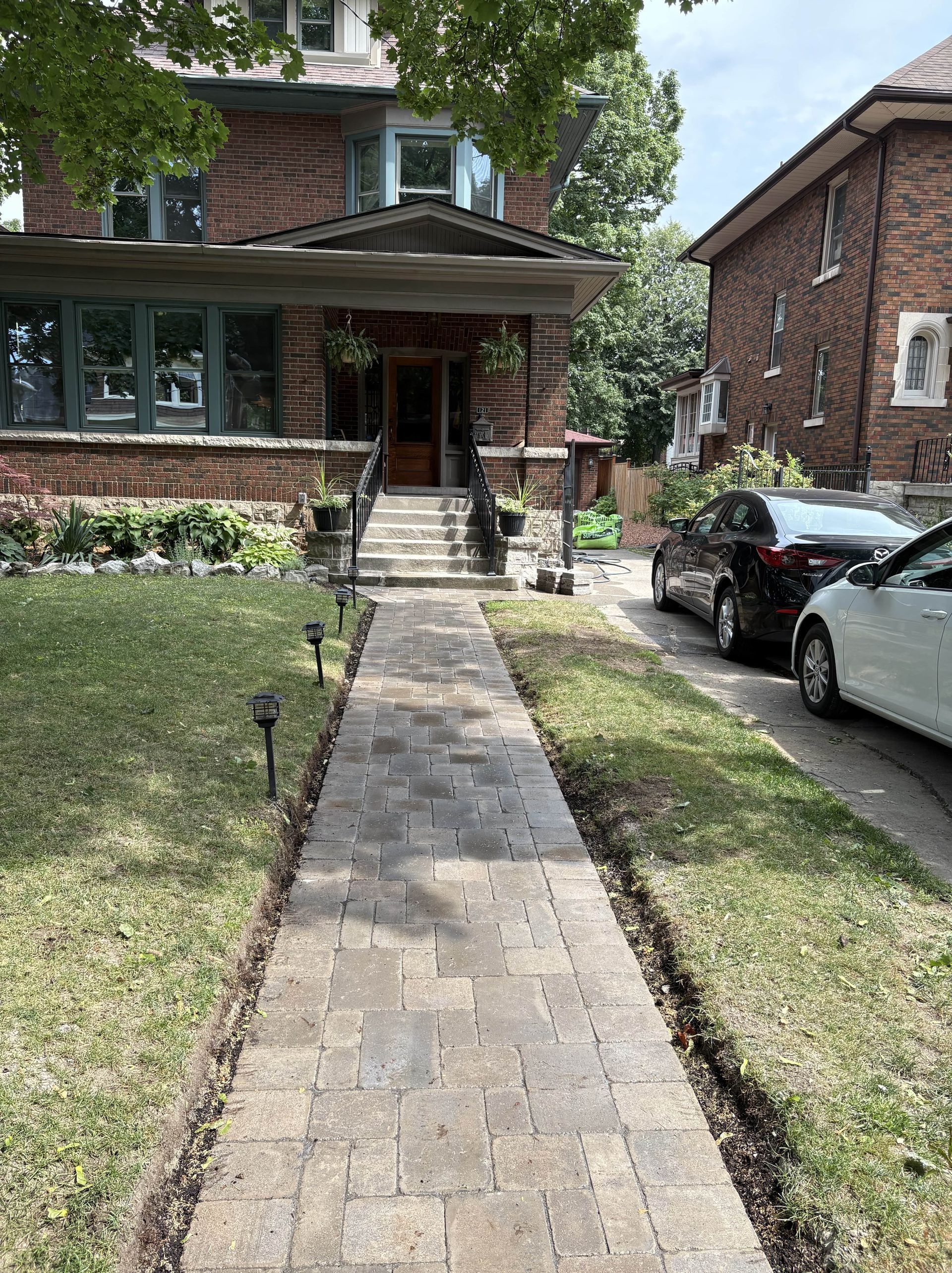 Brick path leading to a brick house with a porch; cars parked along the right.