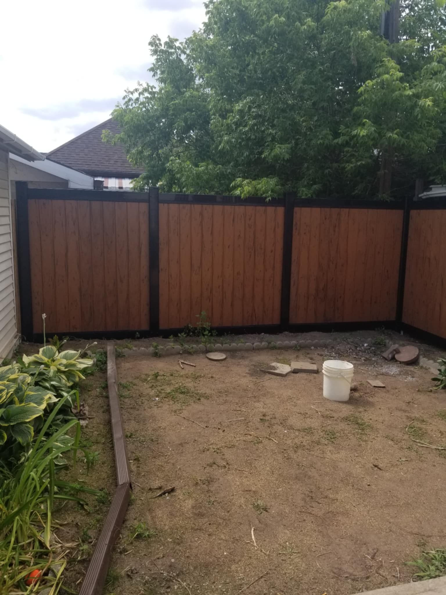 Brown wooden fence in a yard with a patch of grass and a white bucket. Trees in the background.