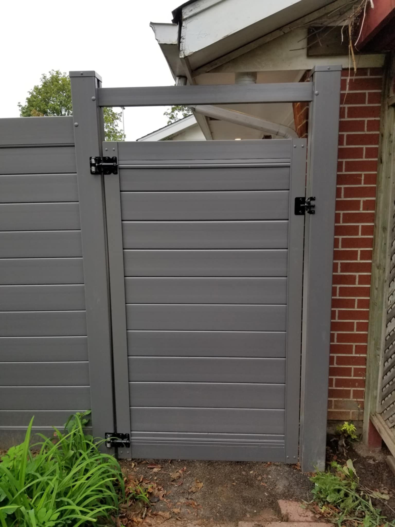 Grey horizontal-slat gate in a gray fence, brick wall to the right, green plants in front.