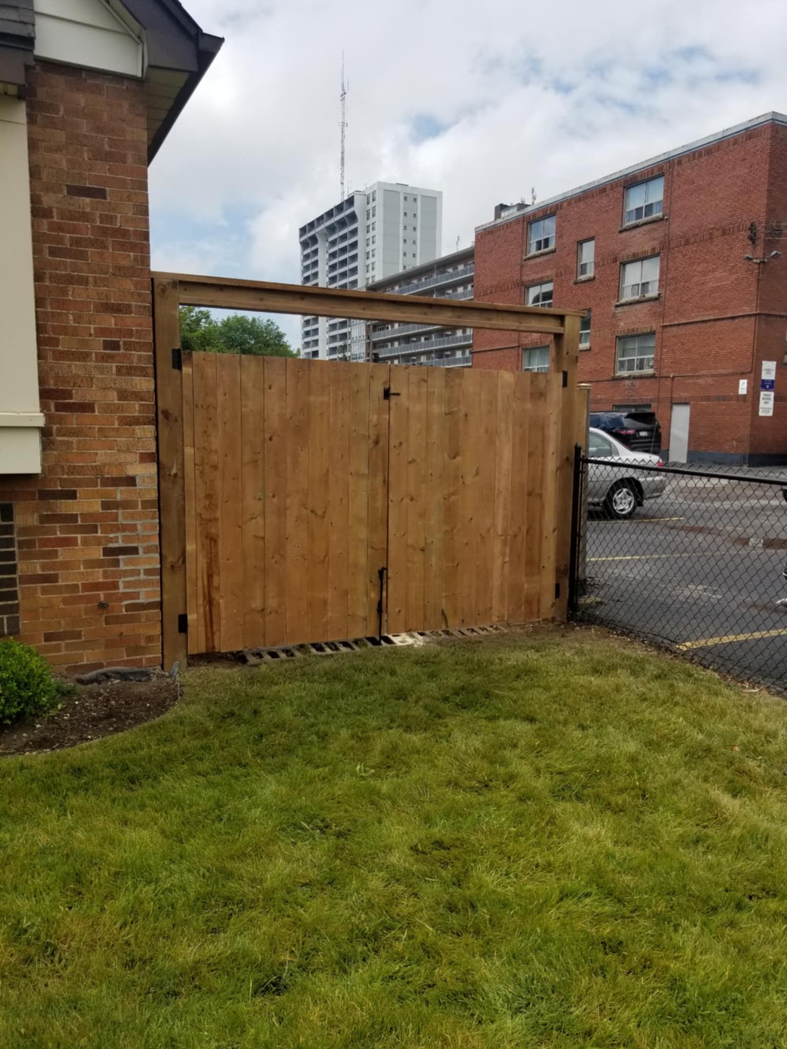 Wooden gate in front of brick building, leading to a parking lot with a gray car. Green grass in front.