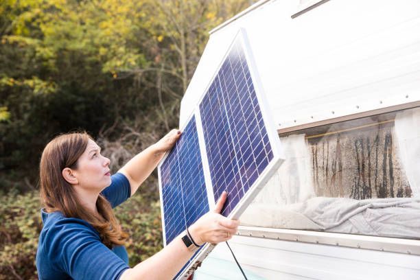 Woman holds a solar panel near a camper in a wooded area, prepping for installation.