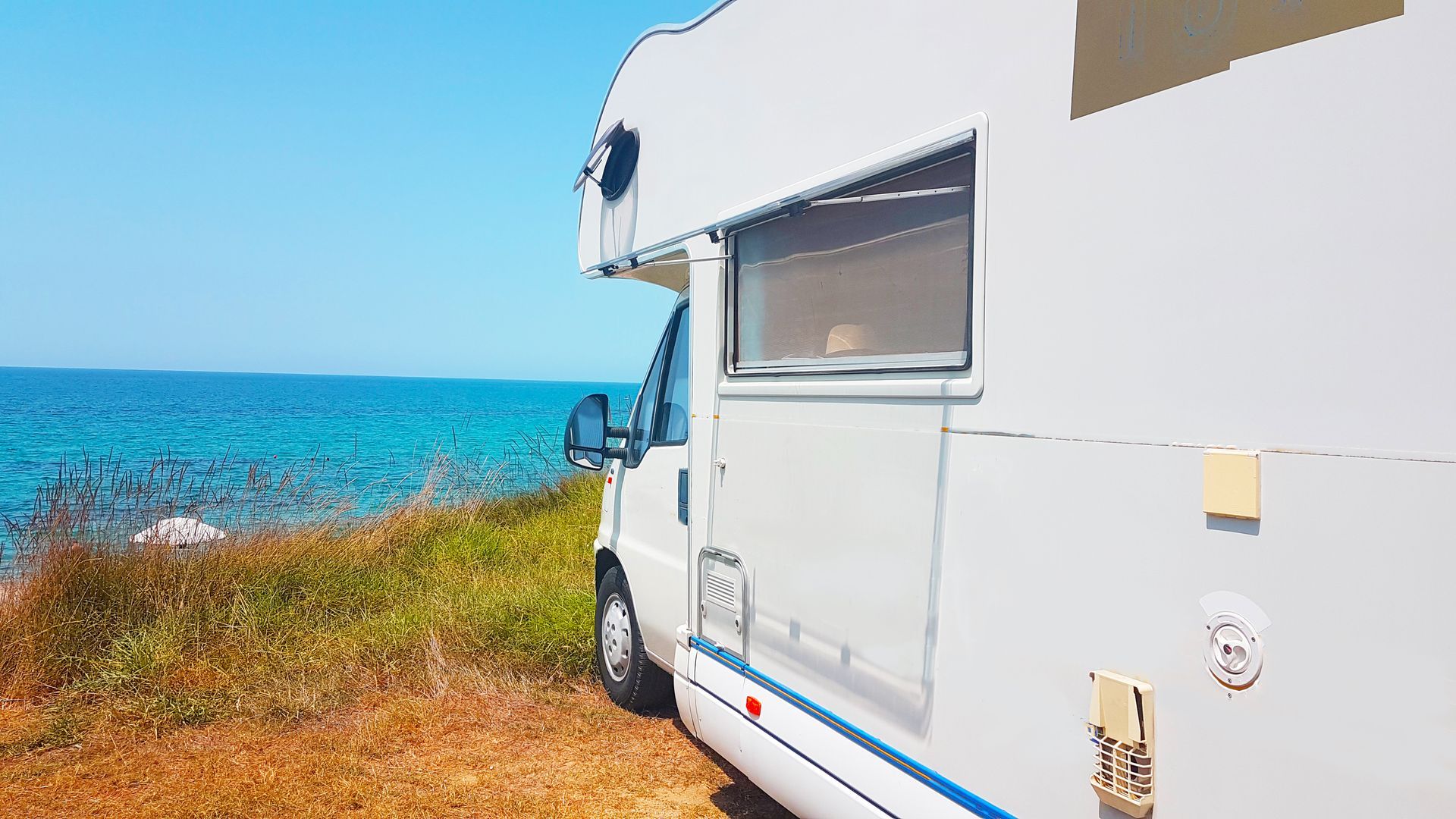 White RV parked near a beach; blue ocean, clear sky in background.