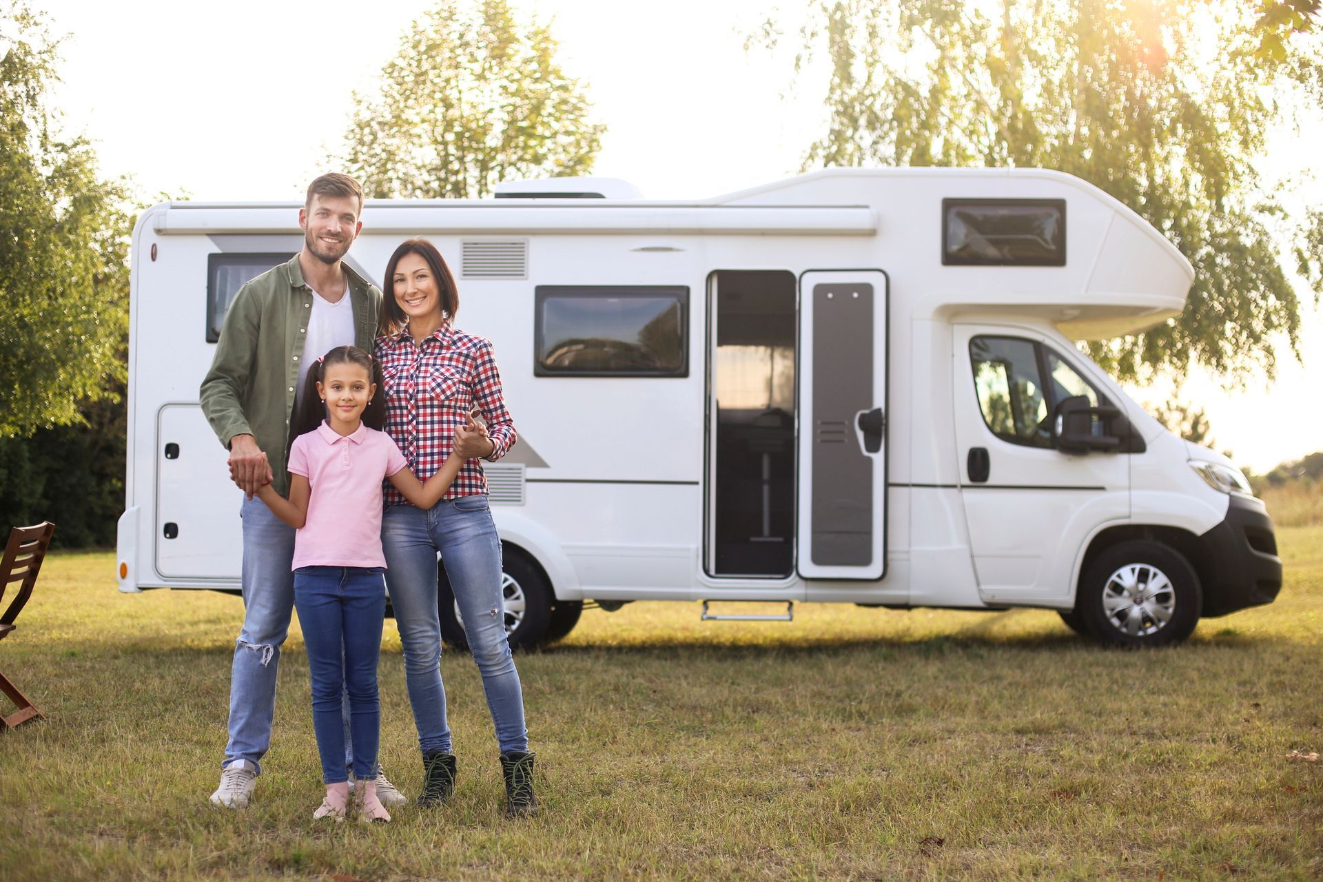 Family of three stands in front of a white RV on a grassy field.