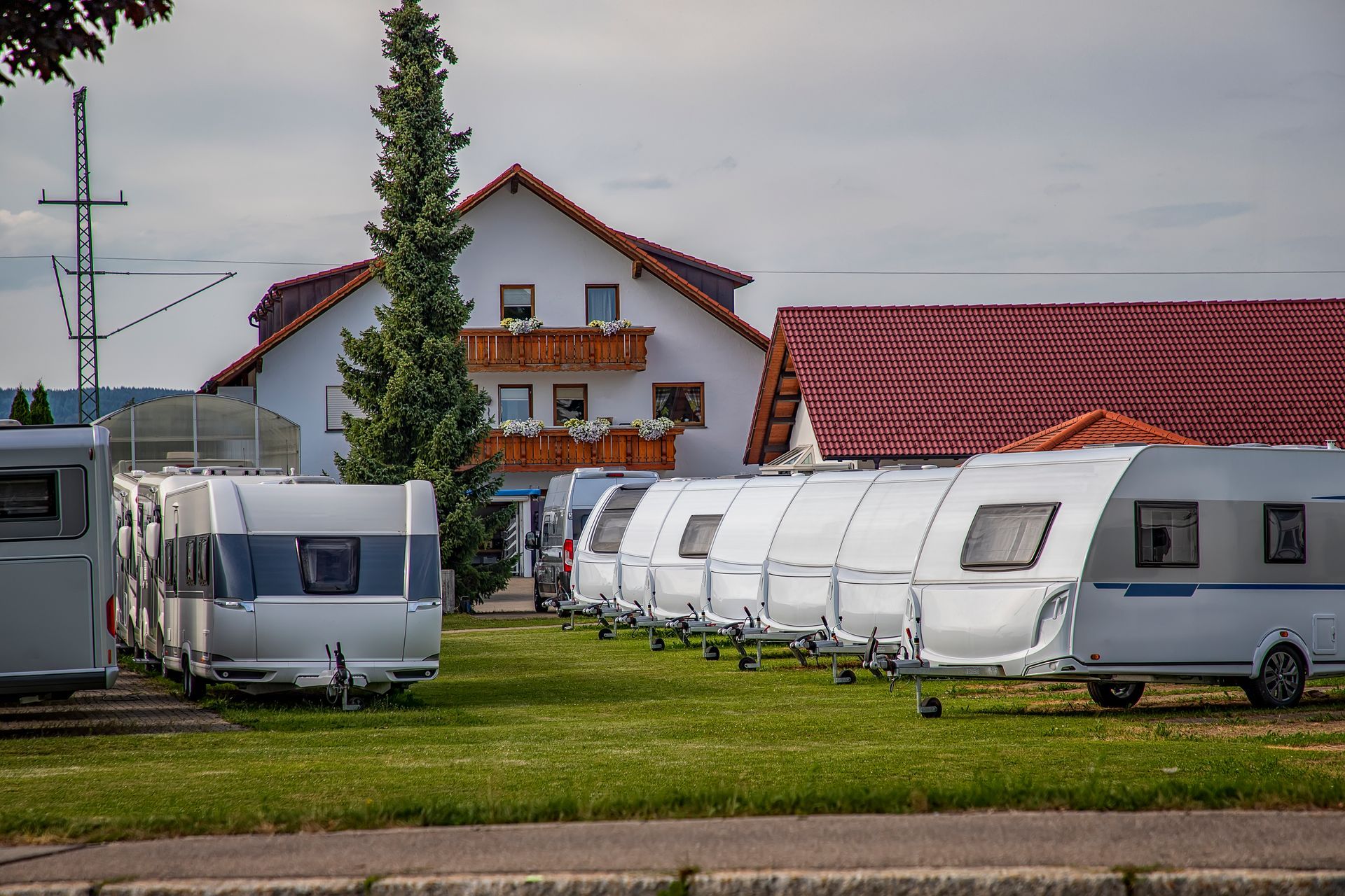 Campers parked on green grass in front of a white building with a red roof and tall pine tree.