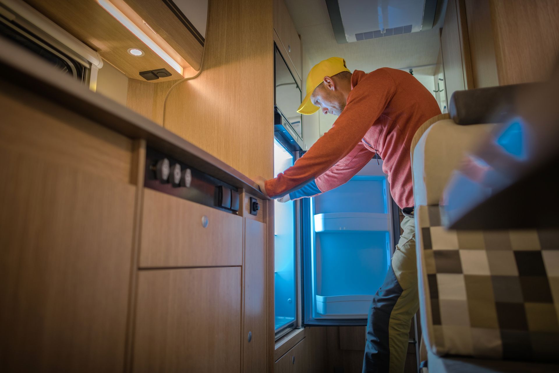 Man in RV opens refrigerator, lit by cool blue light. He wears an orange shirt and yellow cap.
