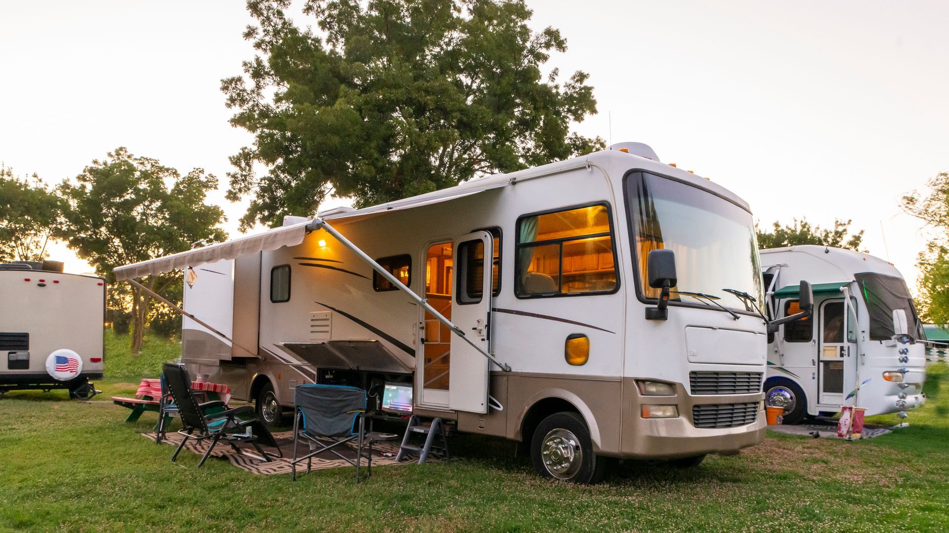 RV parked at a campsite, awning extended, chairs set up; another RV to the right, green grass.