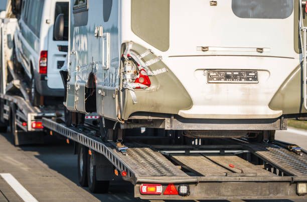 A large trailer transporting a white RV and a smaller white vehicle on a highway.