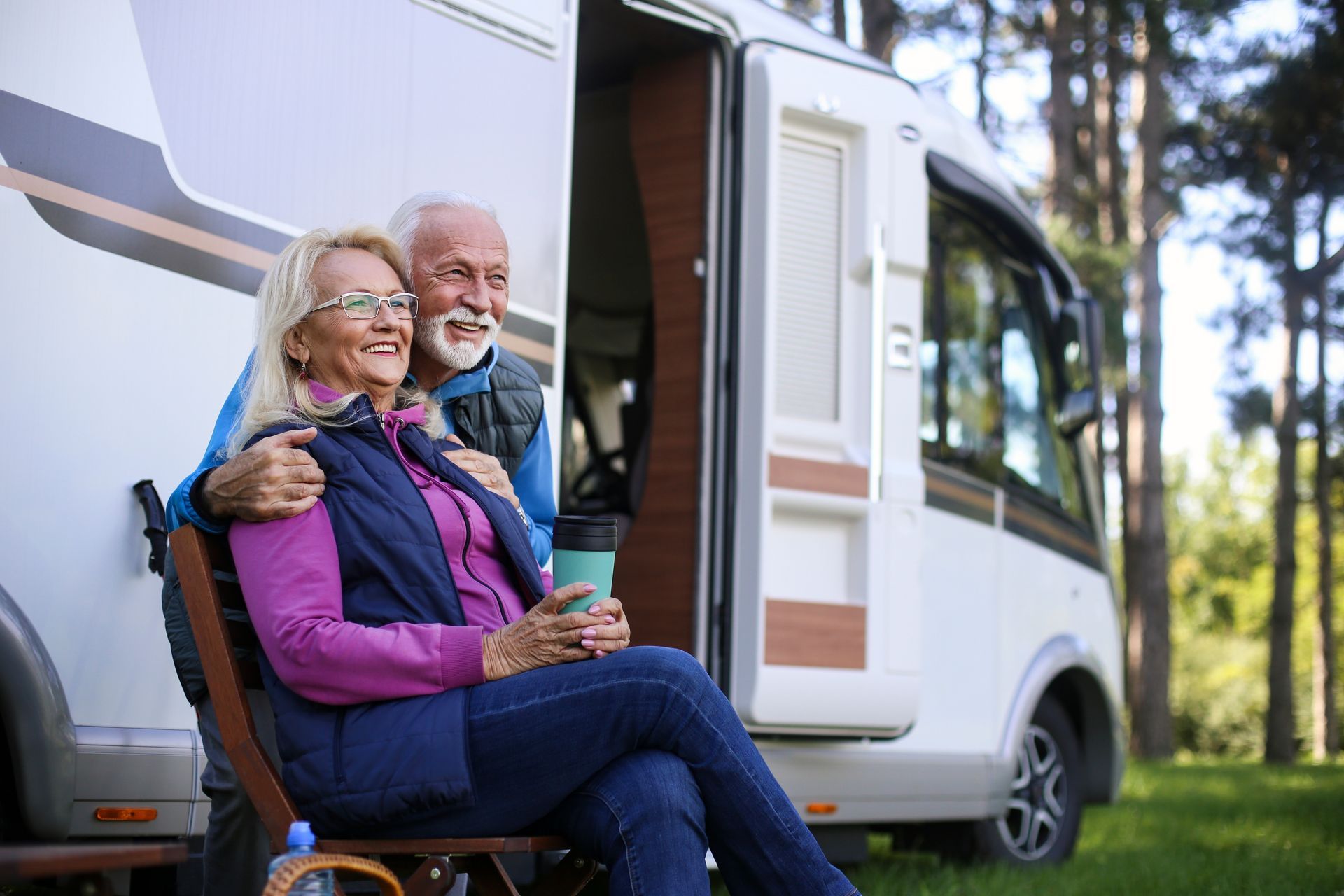Older couple by a RV, woman holding mug, man has arm around her, outdoors.