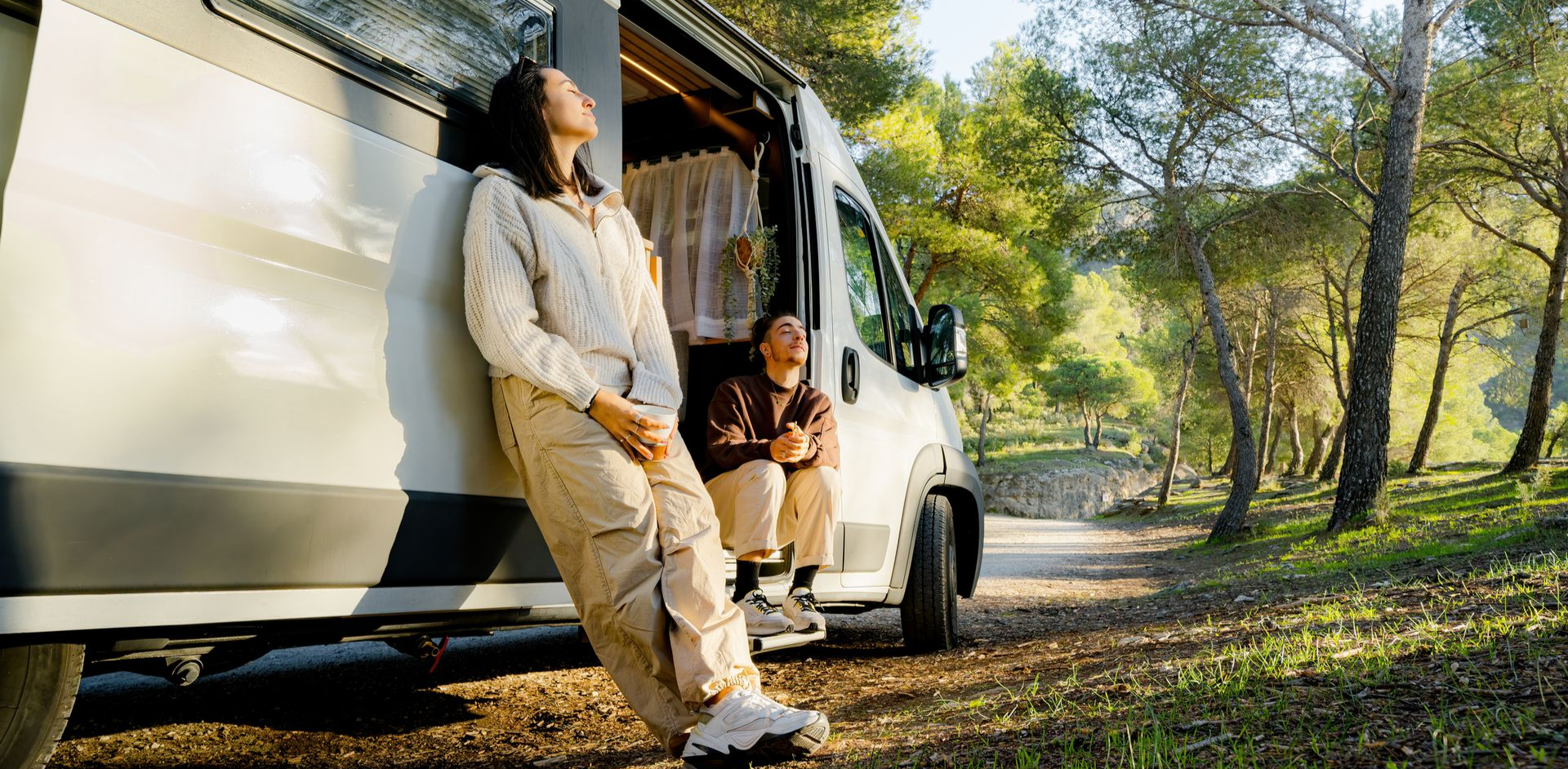 Woman leaning on a van, eyes closed. Man sits in doorway. Sunny, wooded area.