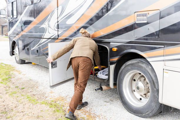 Woman in brown pants opens storage compartment on a black and orange RV parked on gravel.
