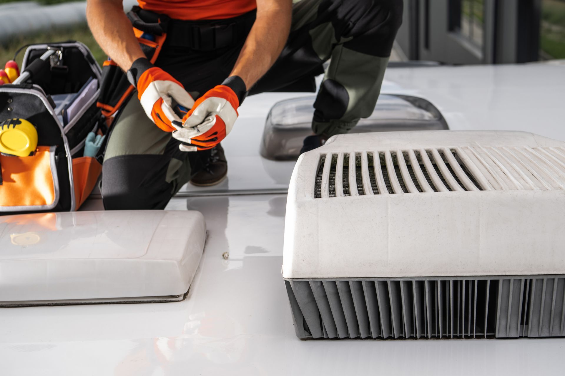 Roofer kneeling on white RV roof, orange gloves, tools, and A/C units.