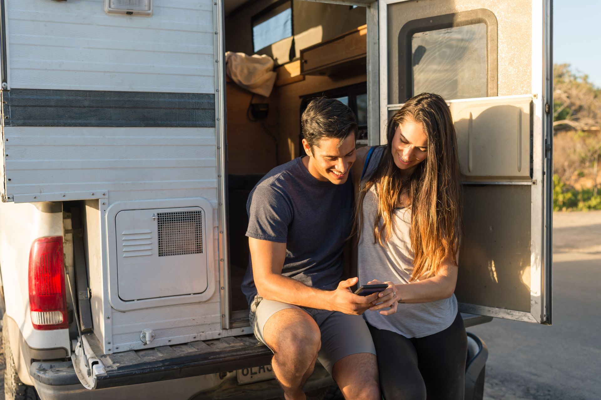 Couple looks at phone, smiling, while sitting on back of campervan with open door; sunny outdoors.