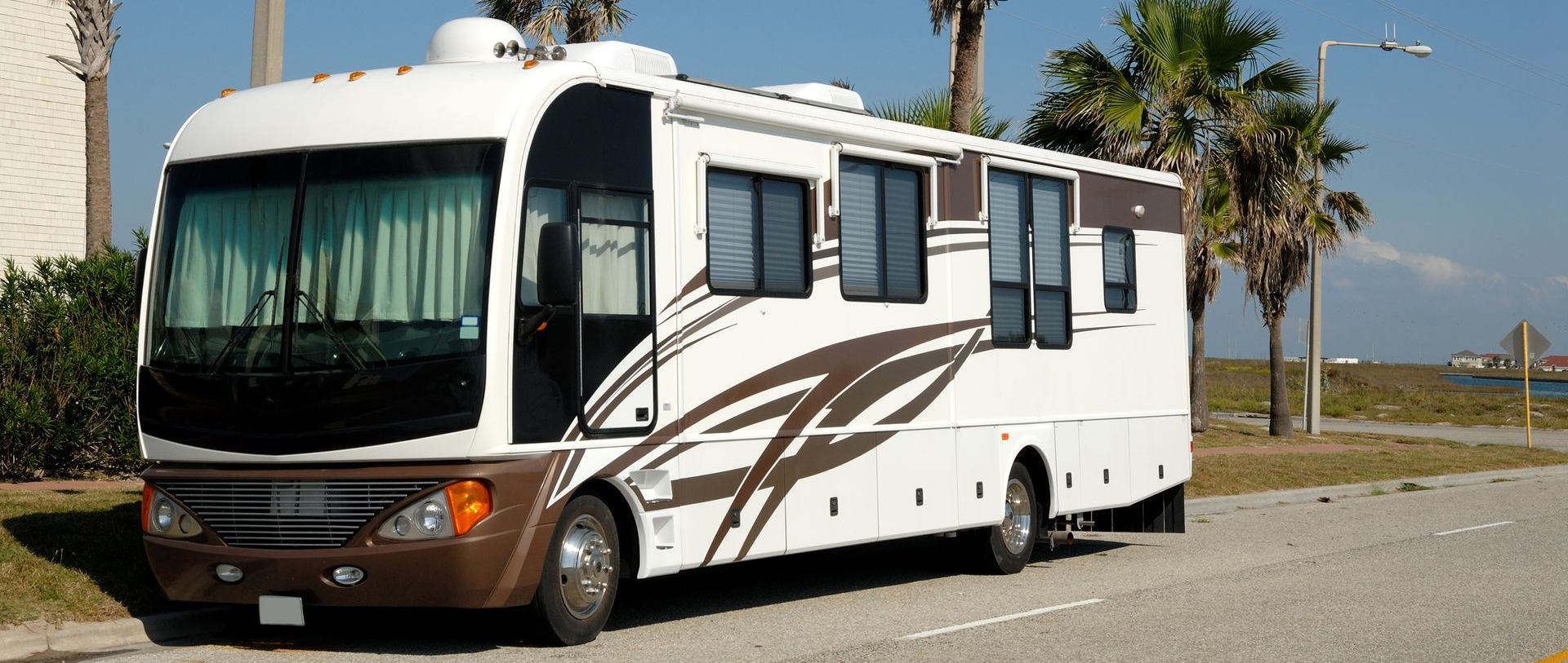 White and brown RV parked on a road near palm trees under a clear blue sky.