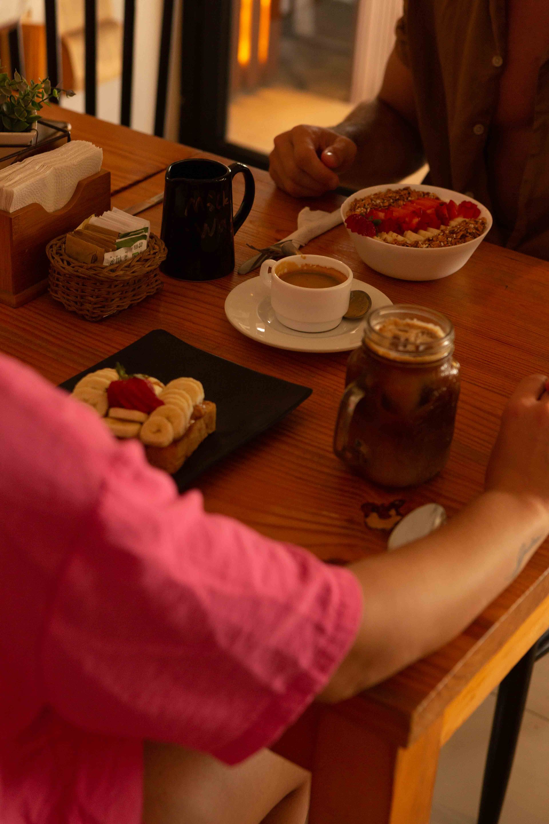 A woman in a pink shirt is sitting at a table with plates of food and a cup of coffee.